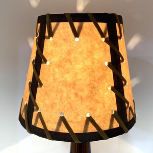 May include: A small, tan lampshade with a dark brown trim and olive green stitching. The lampshade has small holes that emit light. The lamp is on a dark blue base.