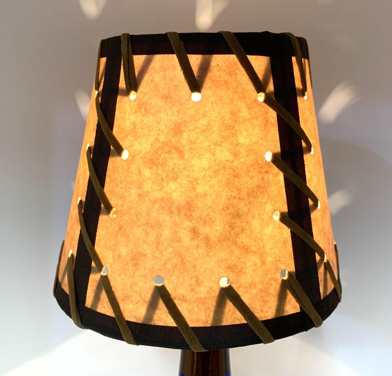 May include: A small, tan lampshade with a dark brown trim and olive green stitching. The lampshade has small holes that emit light. The lamp is on a dark blue base.
