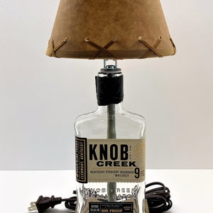 May include: A table lamp made from a repurposed Knob Creek bourbon bottle with a brown leather lampshade. The lamp has a wooden base and a clear glass bottle with the Knob Creek label visible. The lamp is plugged in and ready to use.