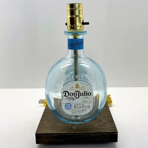 May include: A clear glass bottle lamp with a gold metal lampshade and a wooden base. The bottle has a label that reads "Tequila Don Julio" and "Tequila Blanco".