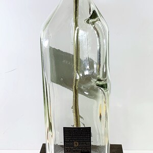 May include: A clear glass bottle lamp with a gold metal lampshade and a brown wooden base. The bottle has a label with the text "DIAGEO" and a barcode. The lamp has a yellow cord and plug.