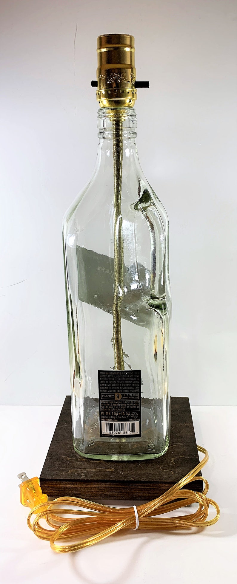 May include: A clear glass bottle lamp with a gold metal lampshade and a brown wooden base. The bottle has a label with the text "DIAGEO" and a barcode. The lamp has a yellow cord and plug.
