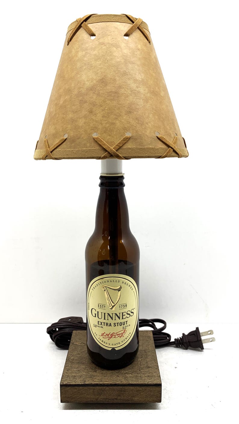 May include: A unique table lamp crafted from a Guinness Extra Stout bottle. It features a tan lampshade with decorative stitching and a wooden base. The lamp includes a power cord and plug.