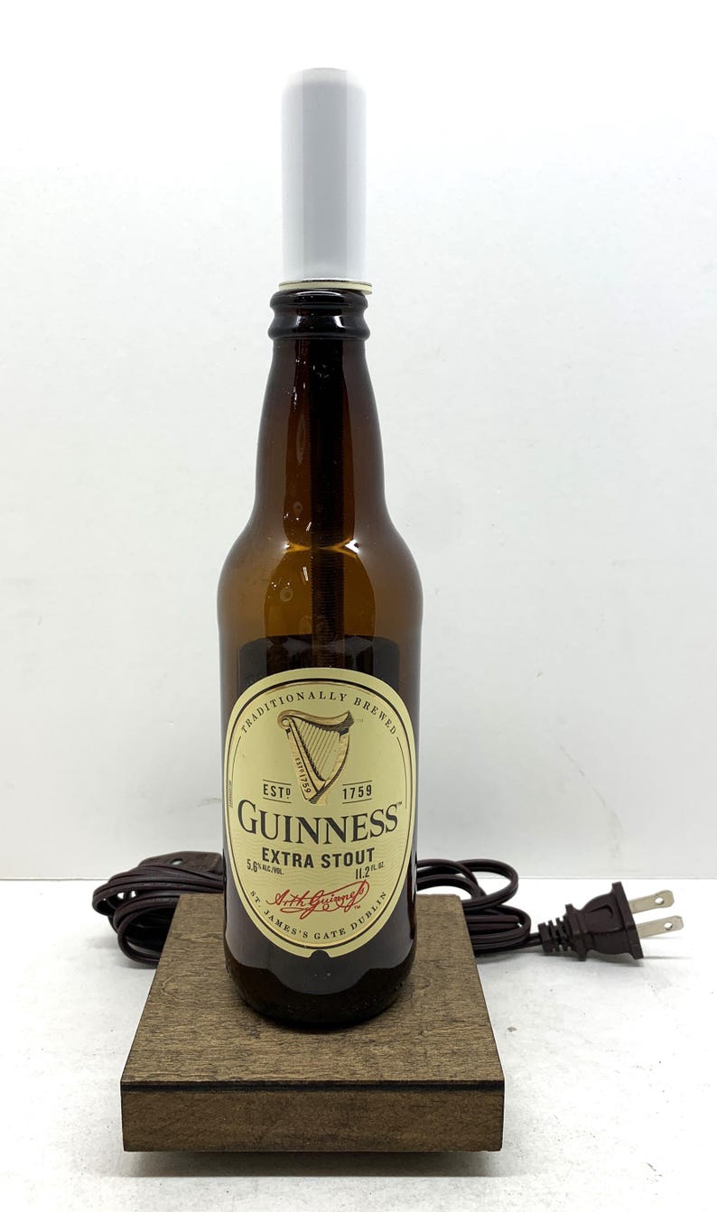May include: A brown Guinness Extra Stout beer bottle with a white cylindrical lamp shade on top. The bottle sits on a small wooden base with a power cord and plug attached. The label reads "Guinness Extra Stout".