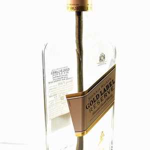 May include: A clear glass bottle lamp with a gold label that reads "Johnnie Walker Gold Label Reserve Blended Scotch Whisky". The bottle is sitting on a dark brown wooden base.