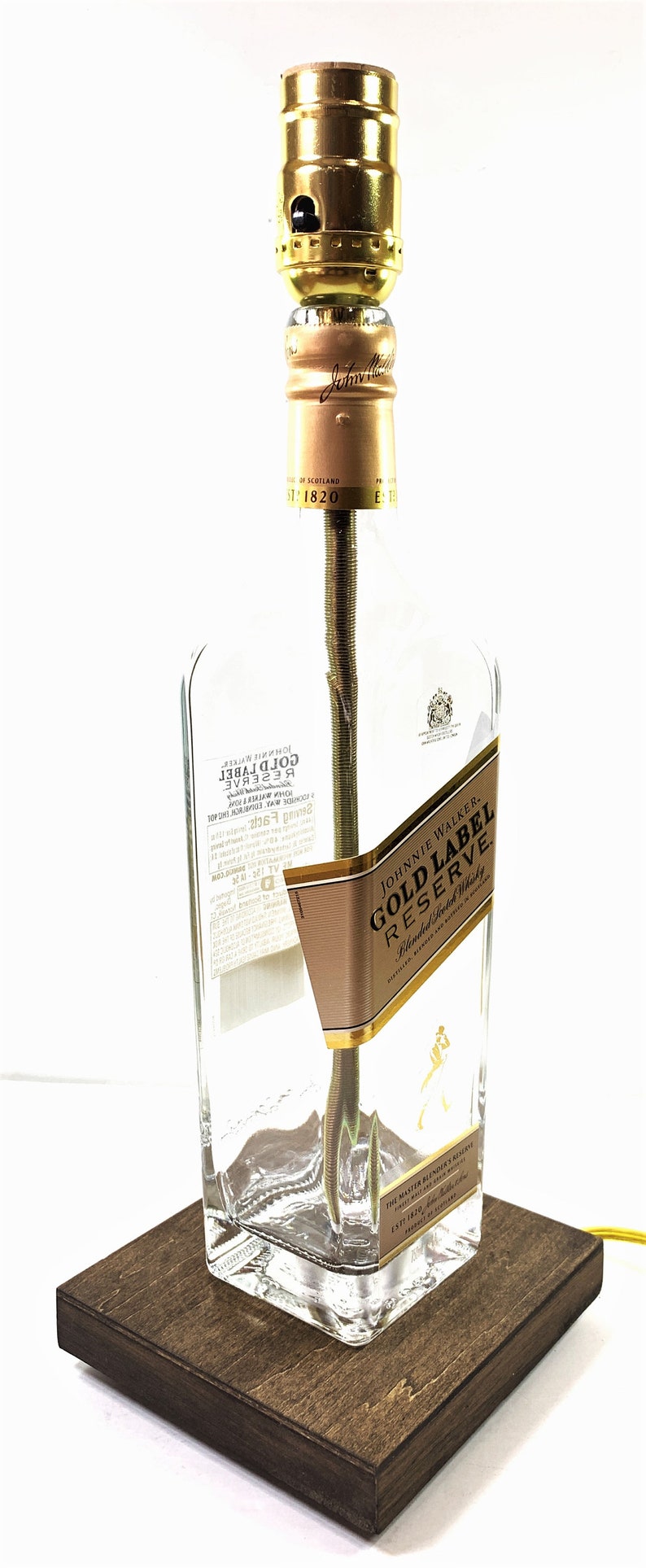 May include: A clear glass bottle lamp with a gold label that reads "Johnnie Walker Gold Label Reserve Blended Scotch Whisky". The bottle is sitting on a dark brown wooden base.