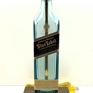K&ouml;nnte beinhalten: Eine umfunktionierte Lampe aus einer Johnnie Walker Blue Label Whiskyflasche. Die hellblaue Glasflasche hat ein schwarz-goldenes Etikett. Die Lampe hat eine goldfarbene Fassung und einen braunen Holzsockel. Ein gelbes Netzkabel ist angebracht.
