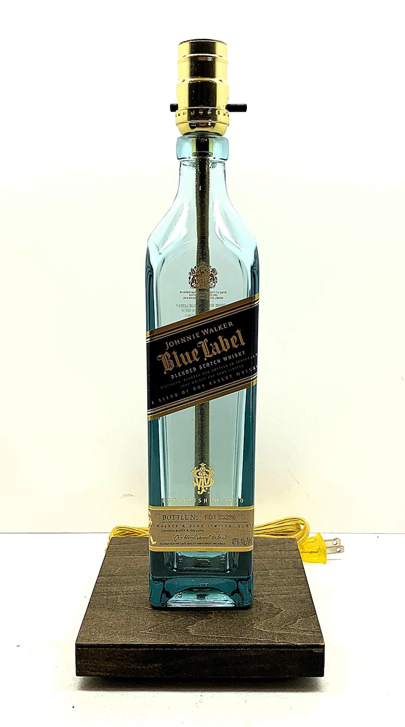 K&ouml;nnte beinhalten: Eine umfunktionierte Lampe aus einer Johnnie Walker Blue Label Whiskyflasche. Die hellblaue Glasflasche hat ein schwarz-goldenes Etikett. Die Lampe hat eine goldfarbene Fassung und einen braunen Holzsockel. Ein gelbes Netzkabel ist angebracht.