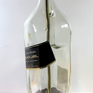 May include: A table lamp made from a repurposed Johnnie Walker Black Label Scotch Whisky bottle. The bottle is clear glass with a black label and gold lettering. The lamp has a gold metal socket and a brown wooden base.