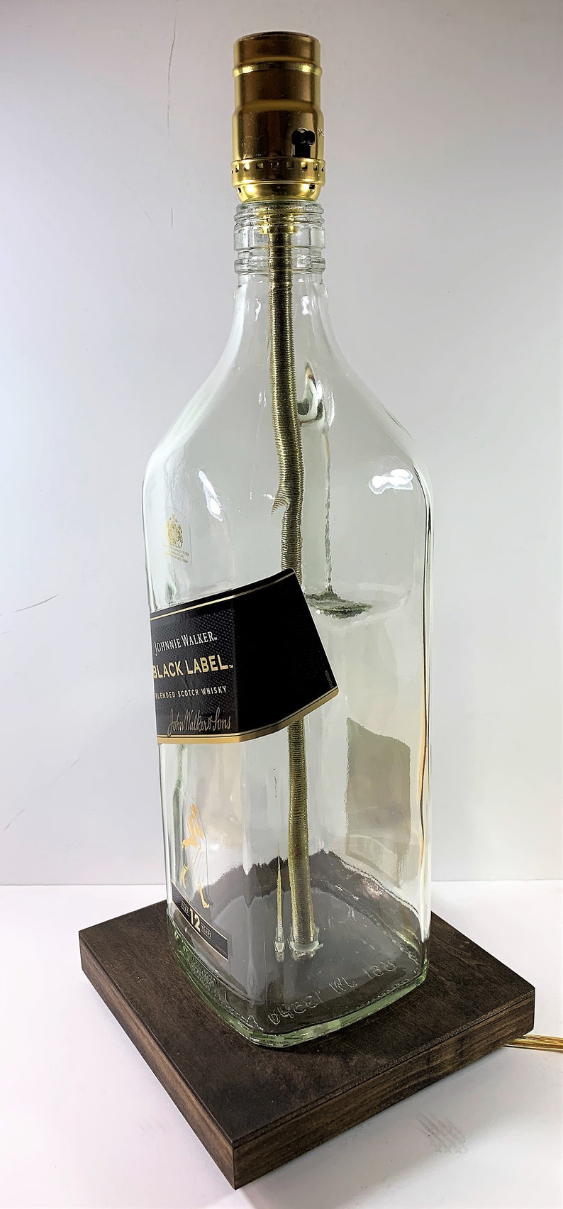 May include: A table lamp made from a repurposed Johnnie Walker Black Label Scotch Whisky bottle. The bottle is clear glass with a black label and gold lettering. The lamp has a gold metal socket and a brown wooden base.