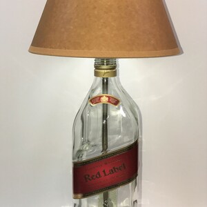 Puede incluir: Una l&aacute;mpara de mesa hecha con una botella de whisky Johnnie Walker Red Label reutilizada con una pantalla marr&oacute;n. La botella es de vidrio transparente con una etiqueta dorada y una base de madera.