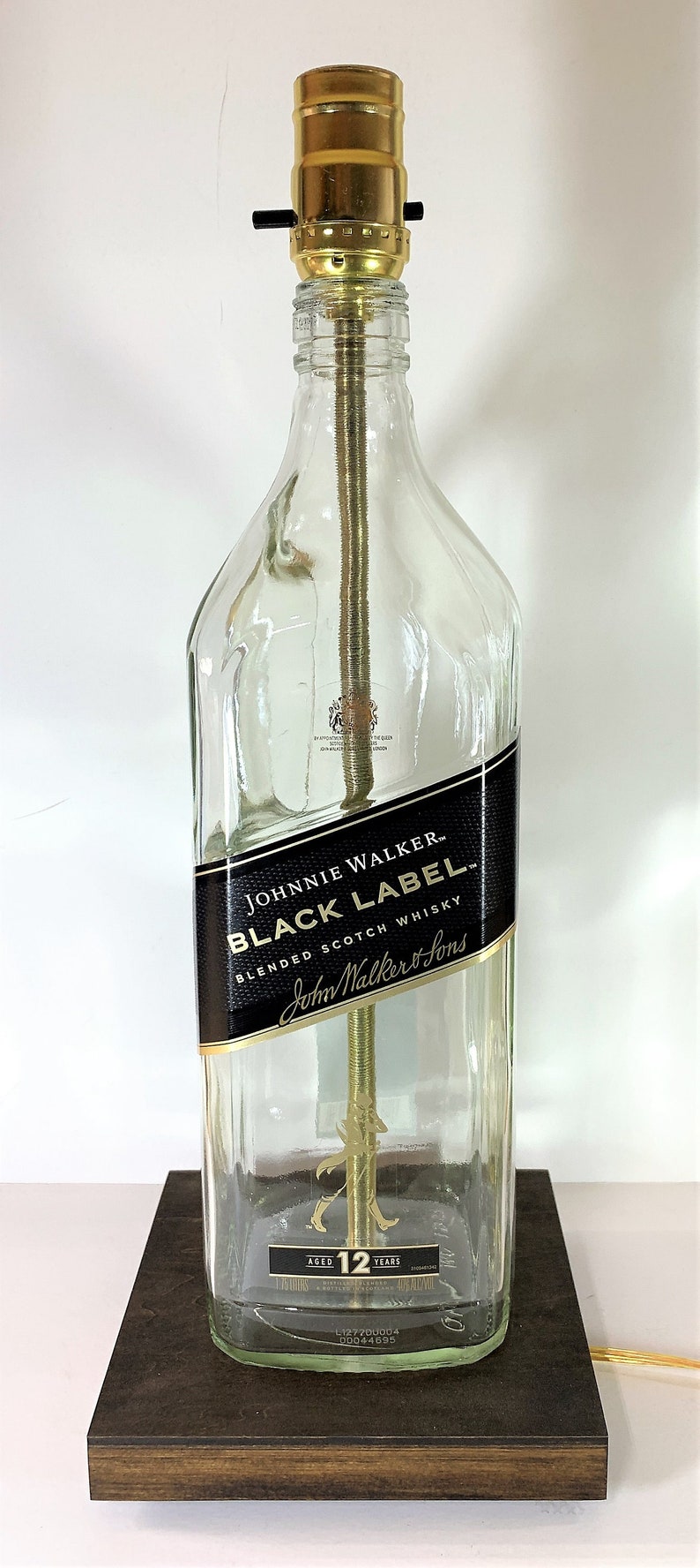 May include: A clear glass bottle lamp with a gold metal base and a black label that reads "Johnnie Walker Black Label Blended Scotch Whisky". The bottle is sitting on a dark brown wooden base.