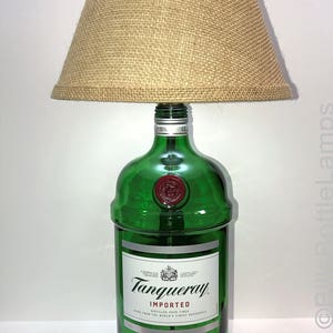 Puede incluir: Una l&aacute;mpara hecha con una botella de ginebra Tanqueray London Dry Gin reutilizada con una pantalla de l&aacute;mpara de arpillera. La botella es de vidrio verde con una etiqueta blanca y las palabras "Tanqueray Importado" y "Ginebra Seca de Londres". La l&aacute;mpara se asienta sobre una base de madera.