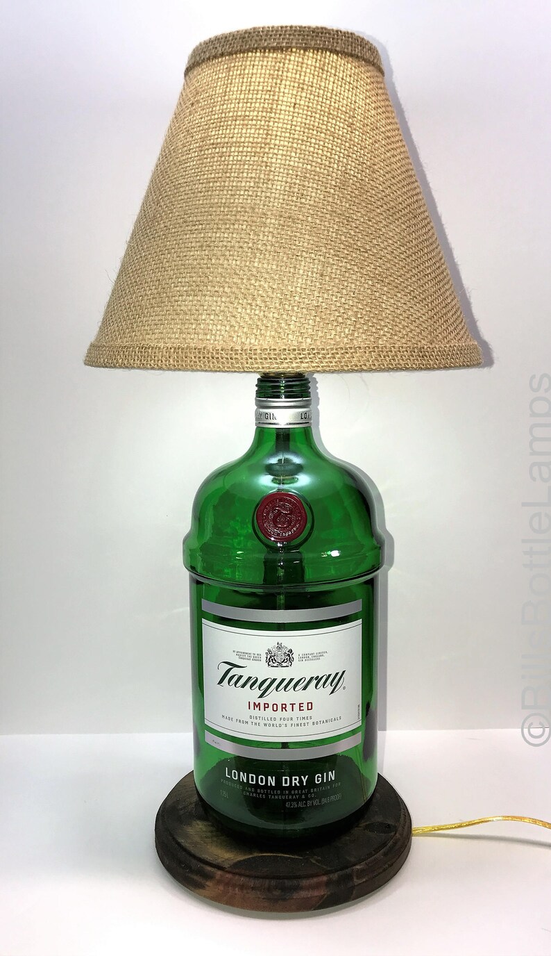 Puede incluir: Una l&aacute;mpara hecha con una botella de ginebra Tanqueray London Dry Gin reutilizada con una pantalla de l&aacute;mpara de arpillera. La botella es de vidrio verde con una etiqueta blanca y las palabras "Tanqueray Importado" y "Ginebra Seca de Londres". La l&aacute;mpara se asienta sobre una base de madera.