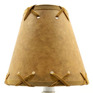 May include: A tan lampshade with a stitched design. The lampshade has a conical shape, with a lighter tan trim and decorative stitching around the top and bottom edges. The stitching is in a cross pattern.