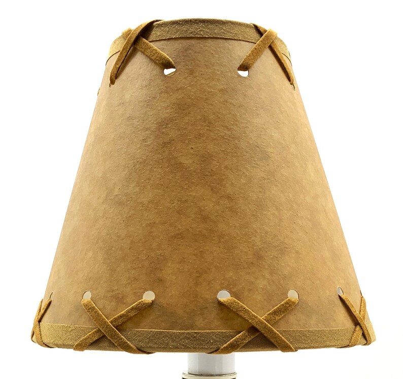 May include: A tan lampshade with a stitched design. The lampshade has a conical shape, with a lighter tan trim and decorative stitching around the top and bottom edges. The stitching is in a cross pattern.