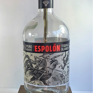May include: A clear glass bottle lamp with a gold metal lampshade. The bottle is labeled "El Espolón Tequila Blanco" with a black and white illustration of a skeleton riding a rooster. The lamp is sitting on a wooden base and has a yellow cord.