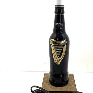 May include: A black Guinness bottle lamp with a white light fixture. The bottle features a gold harp design and the text "GUINNESS DRAUGHT STOUT". The lamp sits on a wooden base with a brown cord and plug.