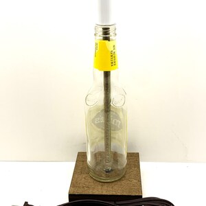 May include: A clear glass bottle lamp with a white bulb and a yellow label. The lamp is mounted on a small, square wooden base. A brown electrical cord and plug are attached to the base. The lamp is a unique home decor item.