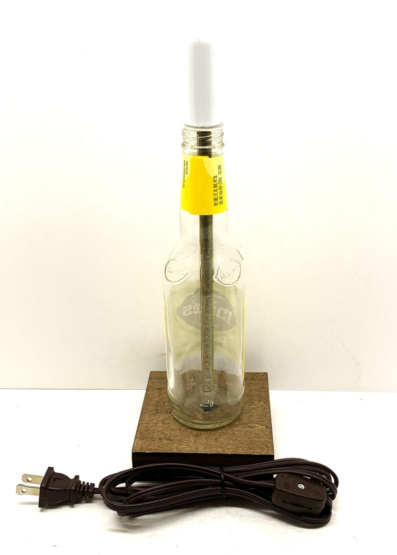May include: A clear glass bottle lamp with a white bulb and a yellow label. The lamp is mounted on a small, square wooden base. A brown electrical cord and plug are attached to the base. The lamp is a unique home decor item.
