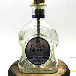 May include: A clear glass bottle lamp with a wooden base. The bottle is labeled "Casa Noble Tequila Crystal Blanco 100% de Agave Azul".