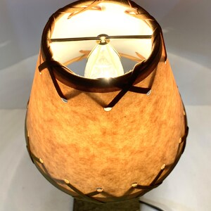 May include: A vintage table lamp with a tan, textured shade and a dark brown trim. The shade is stitched together with dark brown leather-like material. The lamp has a square base and a visible power cord.