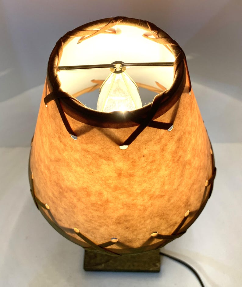 May include: A vintage table lamp with a tan, textured shade and a dark brown trim. The shade is stitched together with dark brown leather-like material. The lamp has a square base and a visible power cord.