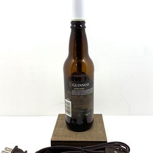 May include: A repurposed Guinness Extra Stout beer bottle lamp. The brown glass bottle has a white light fixture and sits on a wooden base. A brown electrical cord with a plug is attached.