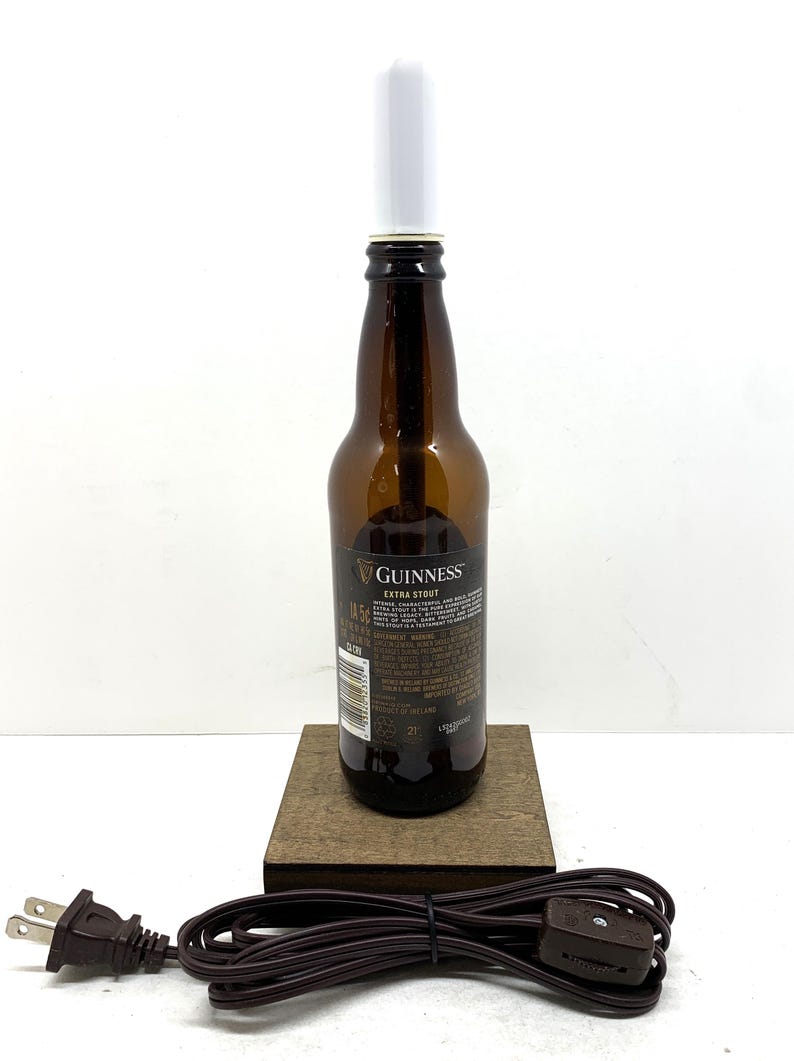 May include: A repurposed Guinness Extra Stout beer bottle lamp. The brown glass bottle has a white light fixture and sits on a wooden base. A brown electrical cord with a plug is attached.
