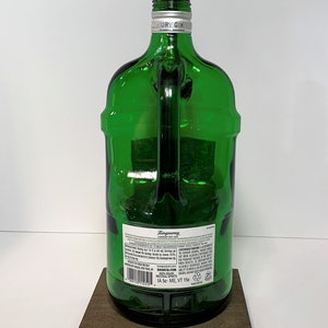 Op de afbeelding: Een groene glazen fleslamp met een gouden fitting en een bruine houten voet. De fles is gelabeld met "Tanqueray London Dry Gin" en heeft een snoer met een gele stekker.