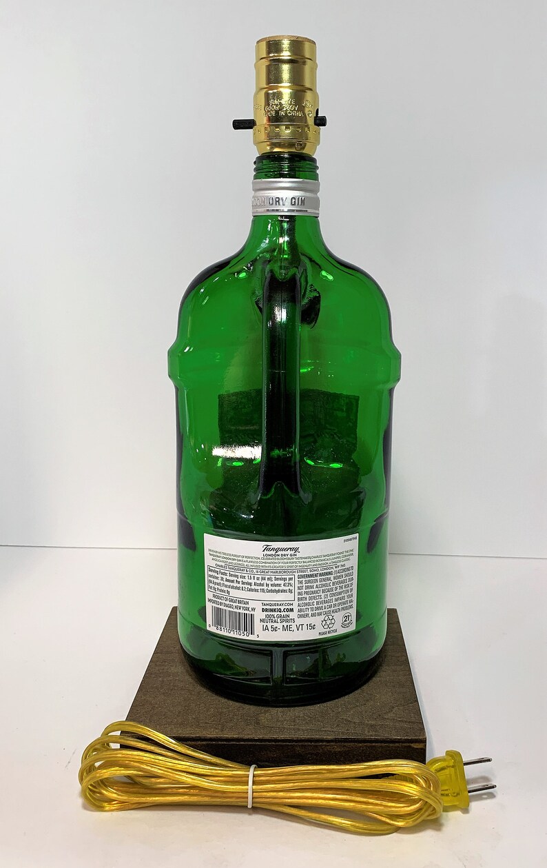 Op de afbeelding: Een groene glazen fleslamp met een gouden fitting en een bruine houten voet. De fles is gelabeld met "Tanqueray London Dry Gin" en heeft een snoer met een gele stekker.