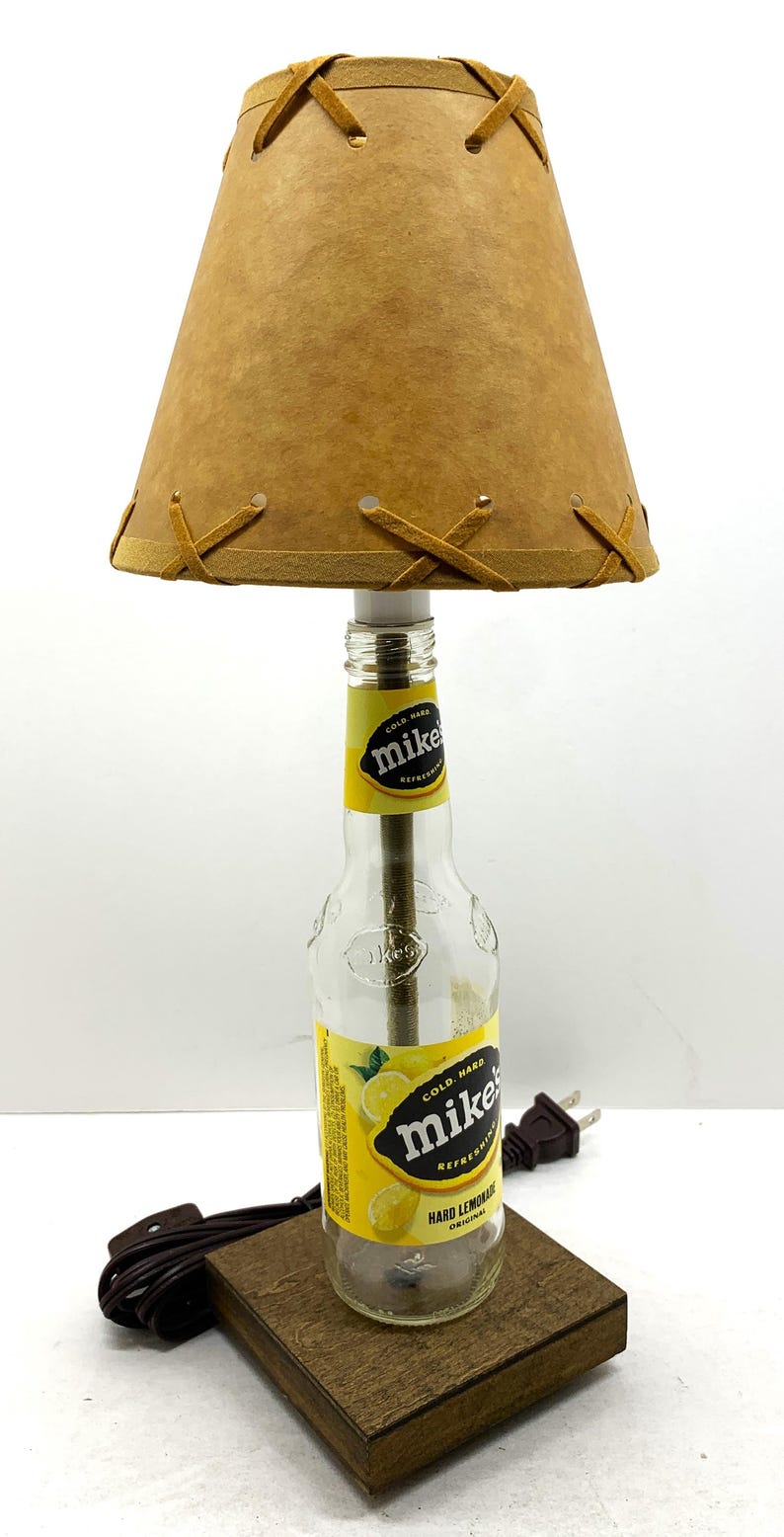May include: A unique table lamp crafted from a repurposed Mike's Hard Lemonade bottle. The lamp features a tan lampshade with decorative stitching and a wooden base. The bottle's label is visible, and the lamp includes a brown power cord.