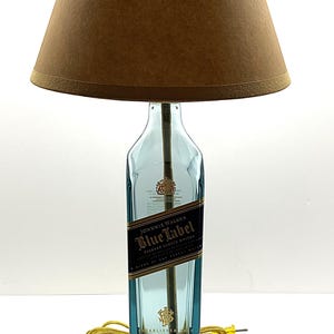 K&ouml;nnte beinhalten: Eine einzigartige Tischlampe aus einer recycelten Johnnie Walker Blue Label Whiskyflasche. Die Lampe hat einen blauen Glasflaschenfu&szlig;, einen braunen Lampenschirm und einen Holzsockel. Die Lampe ist eine dekorative Leuchte.