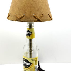 May include: A unique table lamp crafted from a repurposed Mike's Hard Lemonade bottle. It features a tan lampshade with decorative stitching and a wooden base. The lamp includes a power cord and plug.