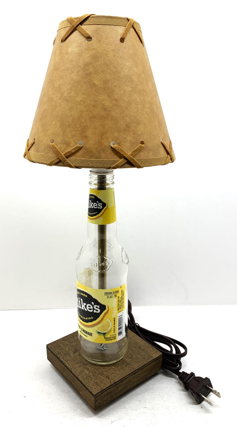 May include: A unique table lamp crafted from a repurposed Mike's Hard Lemonade bottle. It features a tan lampshade with decorative stitching and a wooden base. The lamp includes a power cord and plug.