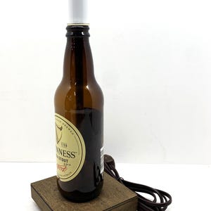 May include: A repurposed Guinness Stout beer bottle lamp. The brown glass bottle sits on a wooden base and has a white cylindrical lamp shade. The lamp includes a brown power cord and plug.