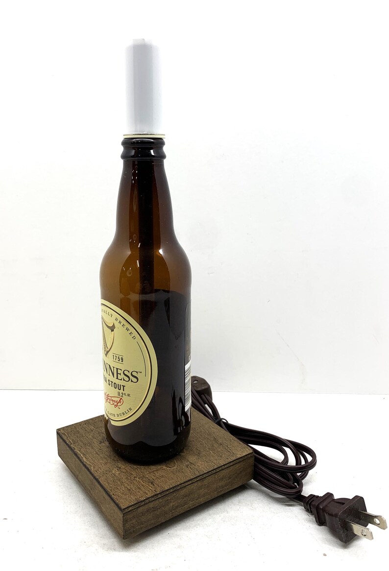 May include: A repurposed Guinness Stout beer bottle lamp. The brown glass bottle sits on a wooden base and has a white cylindrical lamp shade. The lamp includes a brown power cord and plug.