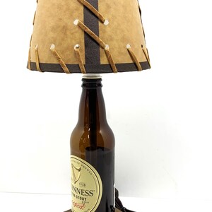 May include: A unique lamp crafted from a Guinness stout beer bottle. The lamp features a tan lampshade with brown accents and decorative stitching. The base is a small, square wooden block. A black power cord and plug are attached.