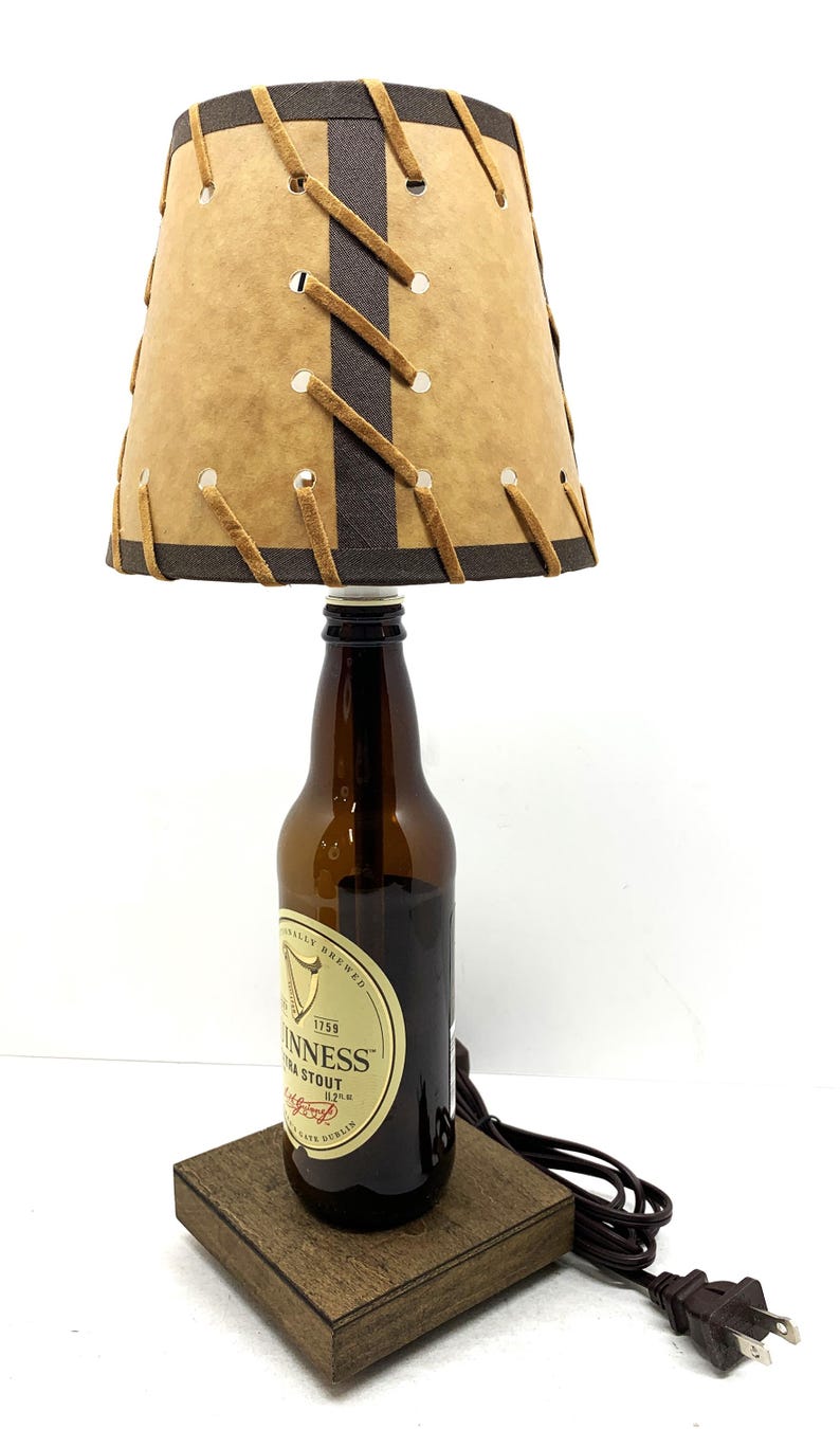 May include: A unique lamp crafted from a Guinness stout beer bottle. The lamp features a tan lampshade with brown accents and decorative stitching. The base is a small, square wooden block. A black power cord and plug are attached.