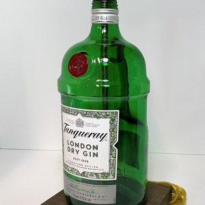 Op de afbeelding: Een groene glazen fleslamp met een gouden metalen lampenkap. De fles is gelabeld met "Tanqueray London Dry Gin Est. 1830". De lamp staat op een donkerbruine houten basis.