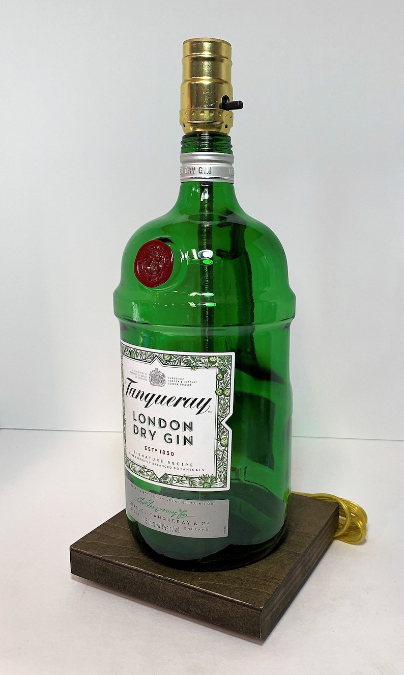 Op de afbeelding: Een groene glazen fleslamp met een gouden metalen lampenkap. De fles is gelabeld met "Tanqueray London Dry Gin Est. 1830". De lamp staat op een donkerbruine houten basis.