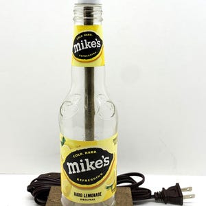 May include: A unique lamp crafted from a Mike's Hard Lemonade bottle. The lamp features a white cylindrical shade, a brown wooden base, and a power cord. The bottle's label reads "Cold. Hard. Mike's Refreshing Hard Lemonade Original."