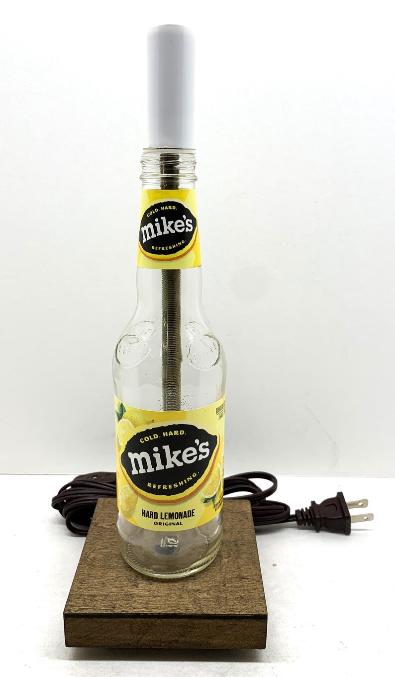 May include: A unique lamp crafted from a Mike's Hard Lemonade bottle. The lamp features a white cylindrical shade, a brown wooden base, and a power cord. The bottle's label reads "Cold. Hard. Mike's Refreshing Hard Lemonade Original."