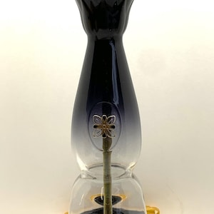 May include: A decorative table lamp with a black and clear glass body, featuring a gold-colored bulb socket and a floral accent. The lamp sits on a dark wooden base and has a yellow power cord.