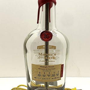 May include: A repurposed Maker's Mark bottle lamp. The clear glass bottle features a red wax seal and a label with text. The lamp has a gold-colored bulb socket and a dark wooden base. A yellow power cord is attached.