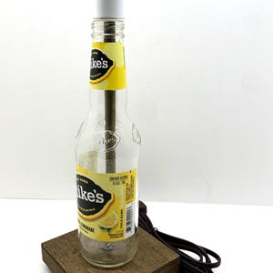 May include: A repurposed lamp made from a clear glass bottle of Mike's Hard Lemonade. The bottle sits on a small, square wooden base and has a white cylindrical lamp shade. The lamp is plugged into a brown electrical cord.