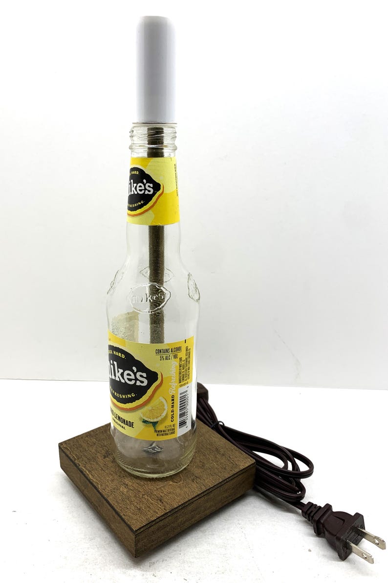 May include: A repurposed lamp made from a clear glass bottle of Mike's Hard Lemonade. The bottle sits on a small, square wooden base and has a white cylindrical lamp shade. The lamp is plugged into a brown electrical cord.