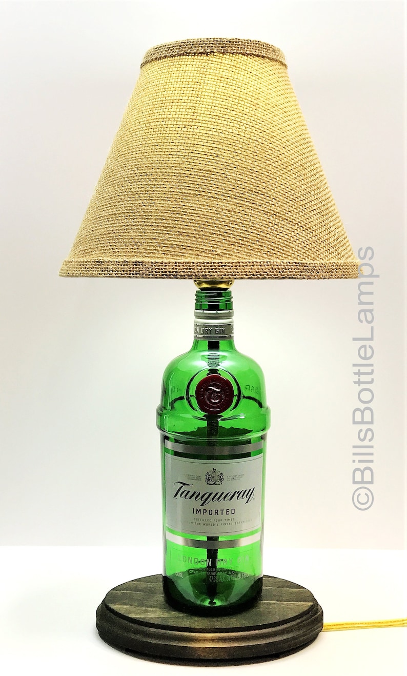 May include: A green glass bottle lamp with a burlap lampshade. The bottle is labeled "Tanqueray Imported" and has a gold crown emblem. The lamp sits on a wooden base.