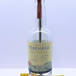 May include: A clear glass bottle lamp with a gold metal lampshade. The bottle is labeled "Teremana Reposado Small Batch Tequila" and has a brown wooden base.