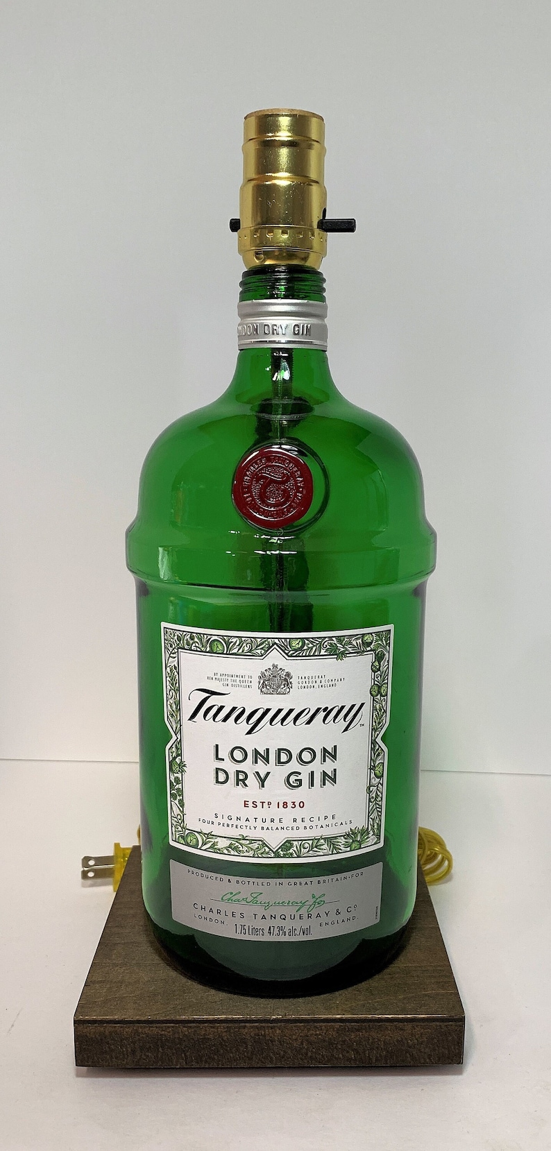 Op de afbeelding: Een groene glazen fles met een gouden bovenkant, gebruikt als lamp. De fles is gelabeld met "Tanqueray London Dry Gin" met de tekst "EST? 1830" en "SIGNATURE RECIPE" op het etiket. De fles staat op een houten voet.